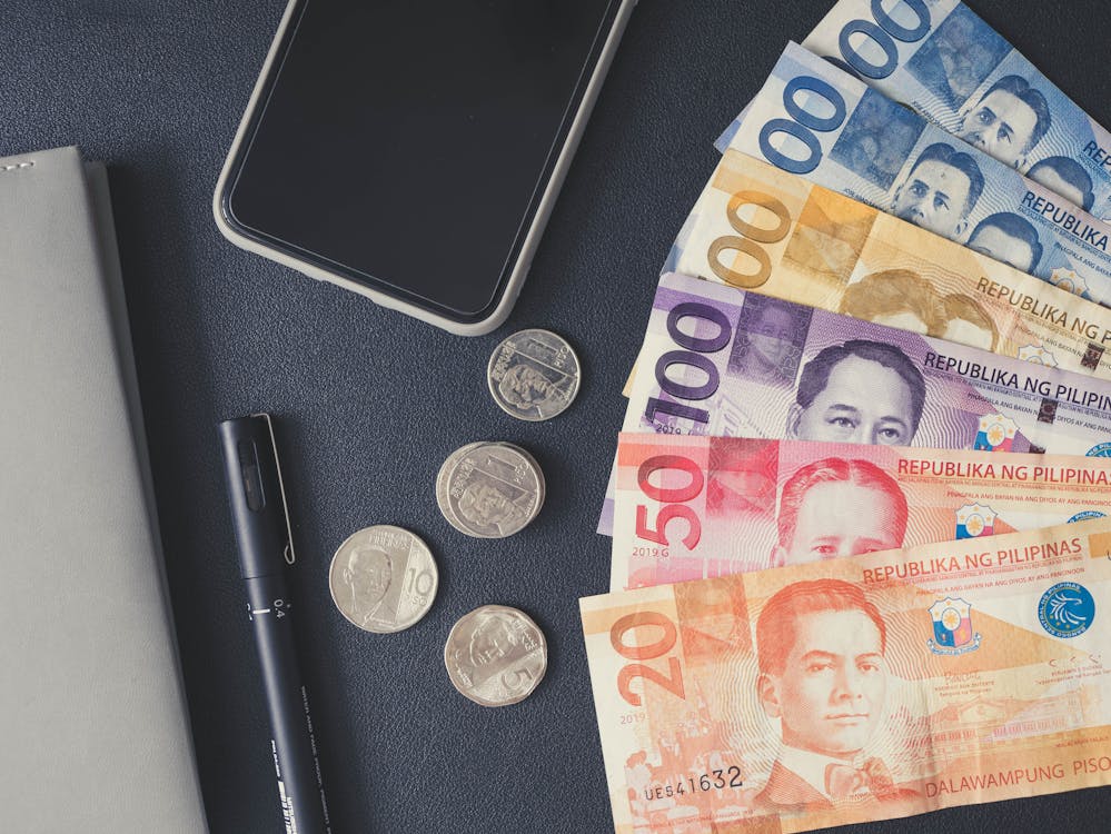 Philippine pesos, smartphone, and pen on a dark surface, representing average salary and financial considerations in the Philippines for outsourcing services.